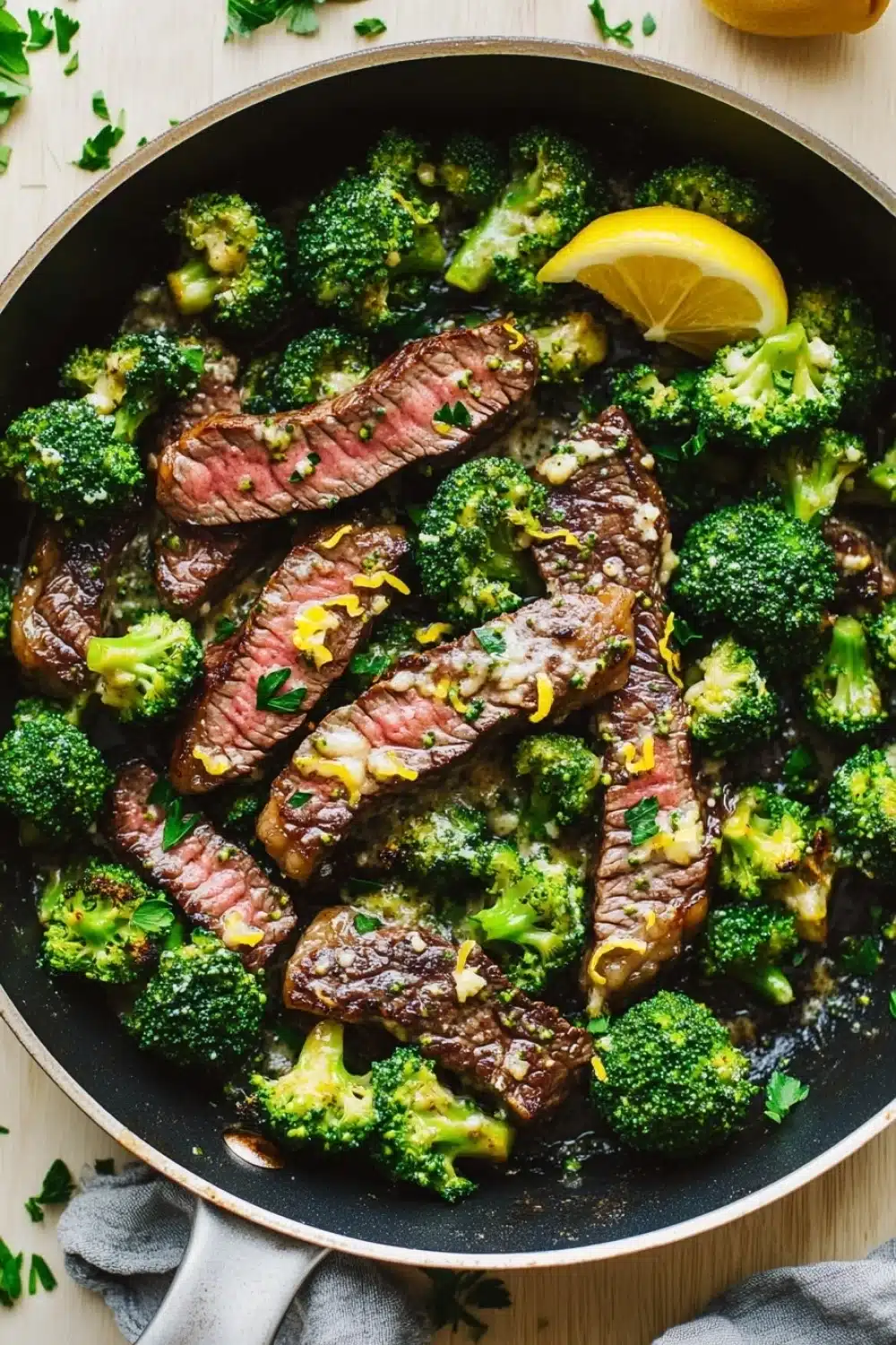 Lemon garlic butter steak and broccoli skillet - the image shows a large black frying pan filled with cooked steak and broccoli. the steak appears to be medium-rare and has a pinkish-brown color. the broccoli is bright green and looks fresh and healthy. there are two lemon wedges on the side of the pan, and a few sprigs of parsley scattered around the pan. the pan is sitting on a wooden table, and there is a gray cloth napkin next to it.