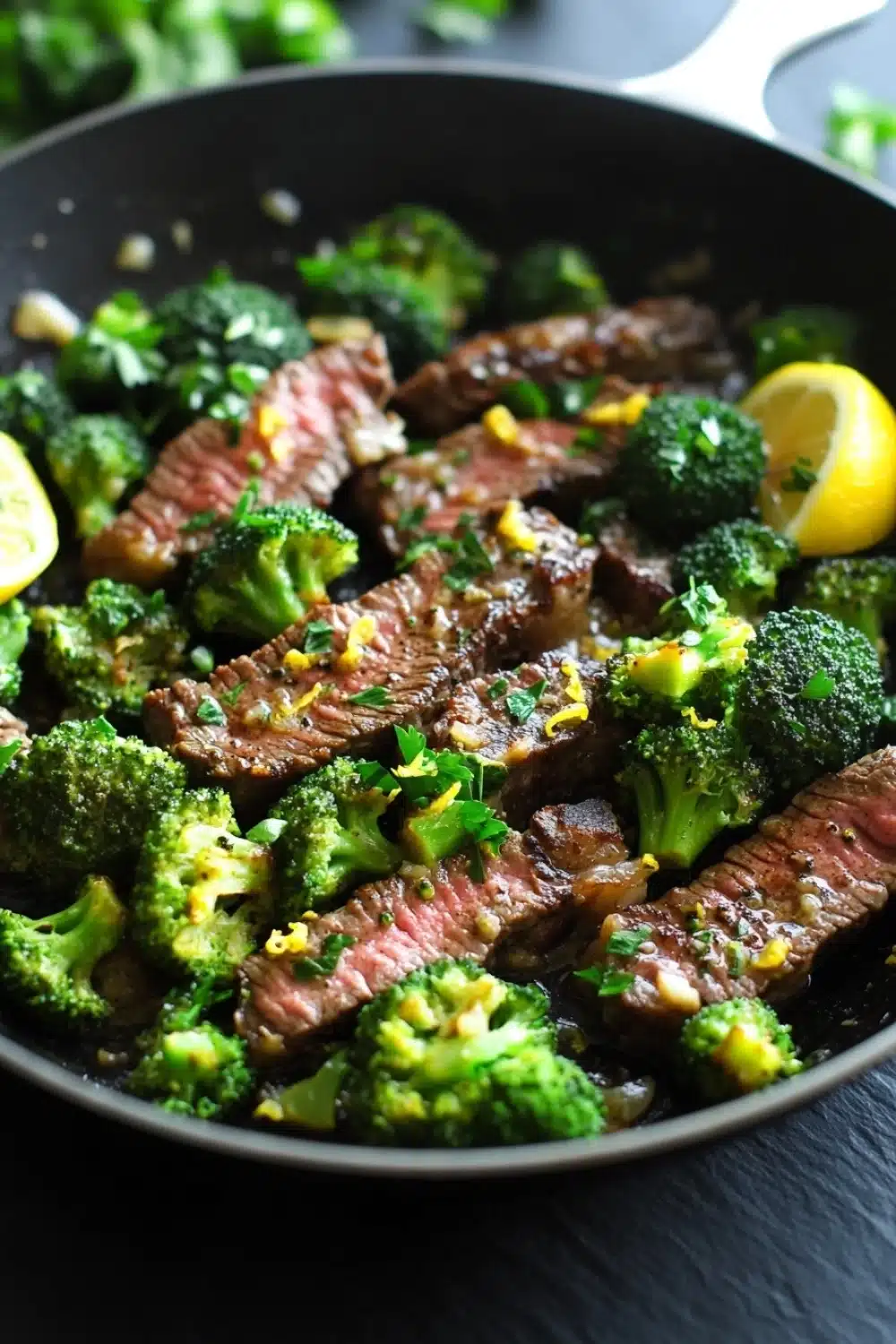 Lemon garlic butter steak and broccoli skillet - the image shows a black frying pan with cooked steak and broccoli in it. the steak appears to be medium-rare and has a pinkish-brown color. the broccoli is bright green and looks fresh and healthy. there are two lemon wedges on either side of the steak, adding a pop of yellow color to the dish. the pan is sitting on a black countertop with a few sprigs of parsley scattered around. the background is blurred, but it seems to be a kitchen countertop.