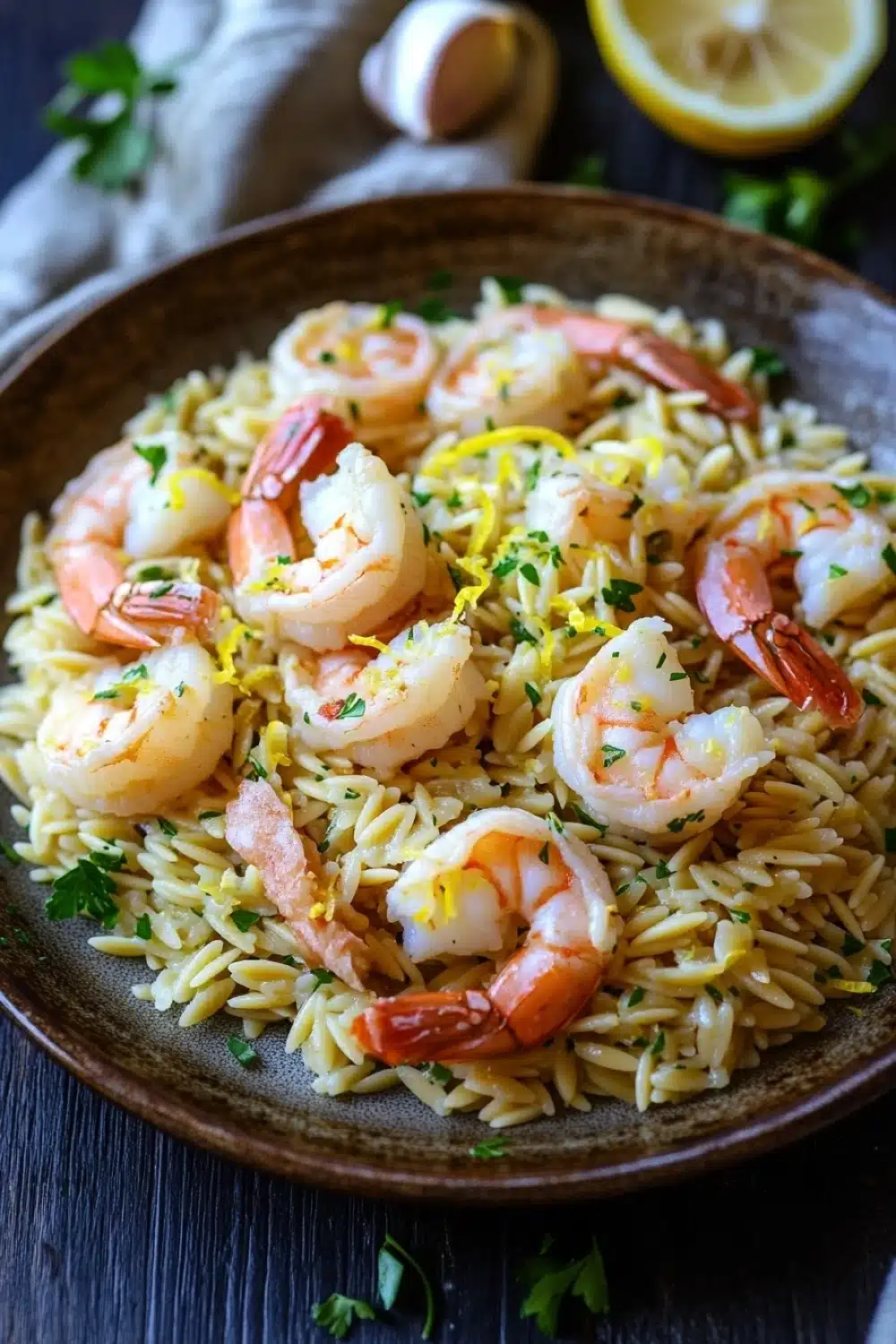 Lemon garlic shrimp orzo skillet - the image shows a plate of pasta dish with shrimp and lemon zest. the dish is served in a brown bowl with a wooden table in the background. the pasta appears to be fusilli, which is a type of spiral-shaped pasta, and is garnished with fresh herbs and lemon wedges. the shrimp are cooked medium-sized and are arranged in a pile on top of the pasta. there is a slice of lemon on the side of the bowl and a few sprigs of parsley scattered around the plate. the overall color scheme of the dish is warm and inviting, with shades of orange, yellow, and green.