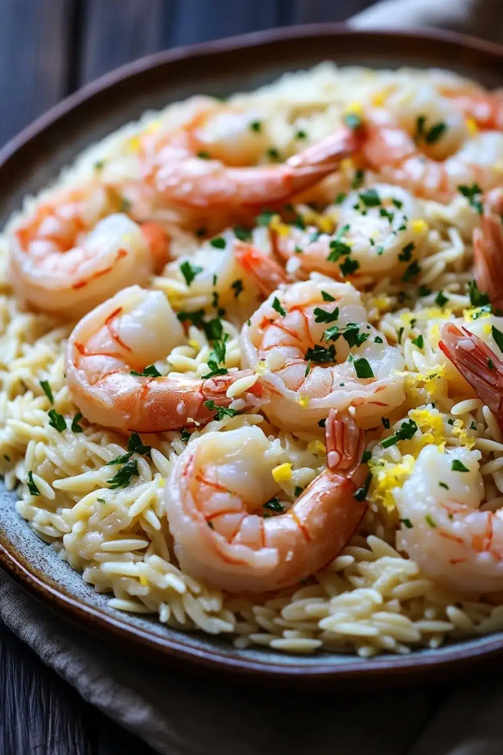 Lemon garlic shrimp orzo skillet - the image shows a plate of pasta dish with shrimp and lemon zest. the dish is served in a brown ceramic plate with a wooden table in the background. the pasta appears to be fusilli, which is a type of pasta, and is mixed with shrimp, lemon, and herbs. the shrimp are cooked medium-sized and are arranged in a circular pattern on top of the pasta. the herbs are scattered throughout the dish, adding a pop of green color to the dish. the plate is garnished with a sprig of parsley.