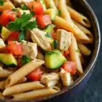 Low calorie chicken pasta - the image is a close-up of a bowl of pasta salad. the bowl is made of dark brown ceramic and is placed on a dark grey countertop. the pasta is long and thin, and it is mixed with diced tomatoes, cucumbers, and chunks of chicken. the salad is garnished with fresh parsley. the colors of the ingredients are vibrant and the dish looks fresh and appetizing.