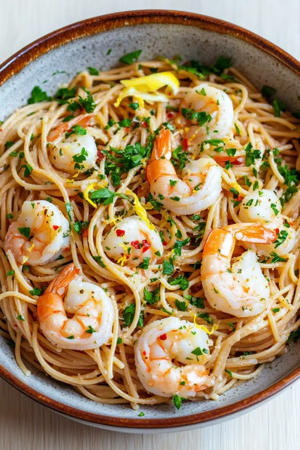 Low calorie shrimp scampi pasta - the image is a close-up of a bowl of pasta dish. the pasta appears to be fettuccine alfredo, with chunks of shrimp scattered throughout. the dish is garnished with chopped parsley and lemon wedges. the bowl is made of ceramic and has a brown rim. the background is a white wooden table.