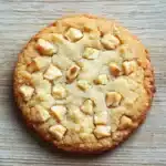 Macadamia nut shortbread cookie - the image is a close-up of a round cookie on a wooden surface. the cookie appears to be freshly baked and has a golden-brown color. it is covered in small chunks of white chocolate chips, which are arranged in a criss-cross pattern on top of the cookie. the chips are evenly distributed throughout the cookie, creating a textured appearance. the background is a light-colored wooden surface, which contrasts with the warm tones of the wood.