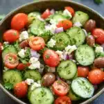 Mediterranean salad cucumber - the image is a close-up of a colorful salad in a large bowl. the salad is made up of sliced cucumbers, cherry tomatoes, red onions, and feta cheese. the cucumbers are cut into thin slices and are arranged in a circular pattern. the tomatoes are bright red and appear to be ripe and juicy. the red onions are scattered throughout the salad, adding a pop of color and texture. the feta is crumbled and adds a creamy texture to the dish. the bowl is sitting on a dark grey surface, and there are a few sprigs of parsley scattered around the bowl.