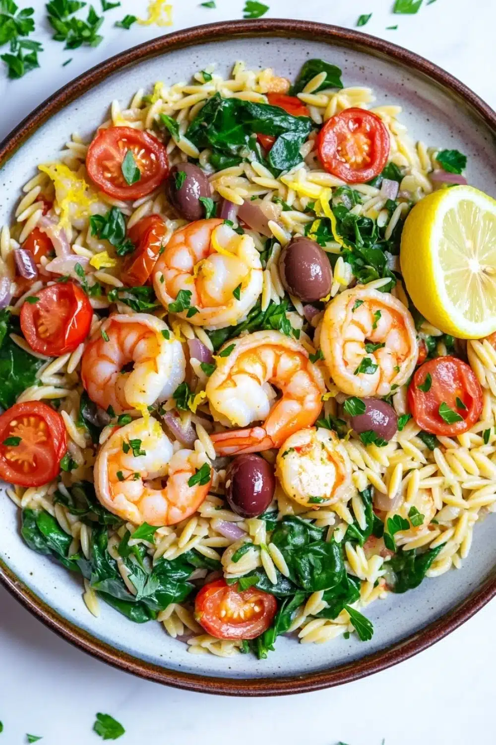 Mediterranean shrimp orzo skillet - the image is a close-up of a plate of pasta salad. the pasta appears to be fusilli, which is a type of italian pasta, and is mixed with various ingredients such as shrimp, cherry tomatoes, olives, and spinach. the dish is garnished with a slice of lemon and a sprig of parsley. the plate is sitting on a white marble countertop. the colors of the ingredients are vibrant and the dish looks fresh and appetizing.