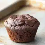 Muffin cacao e yogurt - the image shows a freshly baked chocolate muffin on a baking tray. the muffin is round and has a golden brown color. it appears to be moist and fluffy, with a crumbly texture on top. the tray is lined with parchment paper and there is a dusting of powdered sugar on the surface. the background is blurred, but it seems to be a kitchen countertop.