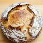 No knead bread new york times - the image is a close-up of a freshly baked loaf of bread on a wooden cutting board. the bread appears to be freshly baked, with a golden brown crust and a soft, fluffy interior. the edges of the bread are slightly frayed, indicating that it has been freshly baked. the loaf is sitting on a light-colored surface, possibly a table or countertop. the background is blurred, making the bread the focal point of the image.