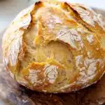 No knead bread with yogurt - the image shows a round loaf of bread on a wooden cutting board. the bread appears to be freshly baked and has a golden brown crust on top. the inside of the bread is covered in a light dusting of powdered sugar, giving it a textured appearance. the cutting board is placed on a white countertop with a small bowl of herbs in the background. the overall mood of the image is rustic and homey.