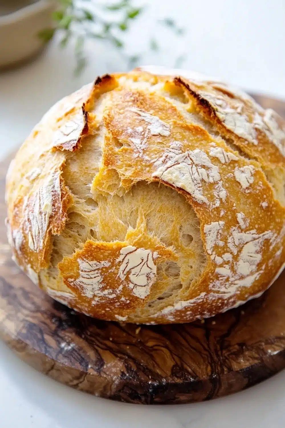 No knead bread with yogurt - the image shows a round loaf of bread on a wooden cutting board. the bread appears to be freshly baked and has a golden brown crust on top. the inside of the bread is covered in a light dusting of powdered sugar, giving it a textured appearance. the cutting board is placed on a white countertop with a small bowl of herbs in the background. the overall mood of the image is rustic and homey.