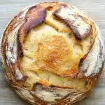 No knead dutch oven bread - the image is a close-up of a round loaf of bread on a wooden surface. the bread appears to be freshly baked and has a golden brown crust on top. the inside of the bread is visible, with a layer of melted cheese oozing out of it. the cheese is a light yellow color and looks like it has been cooked to perfection. the loaf is resting on a light-colored wooden surface, which contrasts nicely with the warm tones of the wood. the overall mood of the image is rustic and cozy.