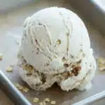 Oatmeal cookie ice cream - the image shows a scoop of vanilla ice cream in a metal baking tray. the ice cream is light brown in color and has a crumbly texture. it is topped with a sprinkle of chopped nuts, giving it a crunchy appearance. the tray is sitting on a wooden surface, and the background is blurred, making the ice cream the focal point of the image.