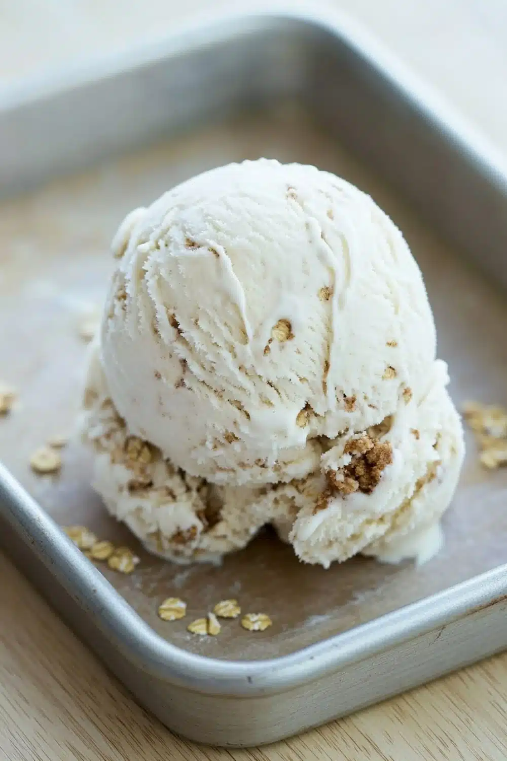 Oatmeal cookie ice cream - the image shows a scoop of vanilla ice cream in a metal baking tray. the ice cream is light brown in color and has a crumbly texture. it is topped with a sprinkle of chopped nuts, giving it a crunchy appearance. the tray is sitting on a wooden surface, and the background is blurred, making the ice cream the focal point of the image.