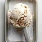 Oatmeal cookie ice cream - the image shows a scoop of vanilla ice cream in a square metal container. the ice cream is topped with chopped nuts, giving it a crunchy texture. the container is sitting on a white marble countertop. a silver ice cream stick is resting on the side of the container.