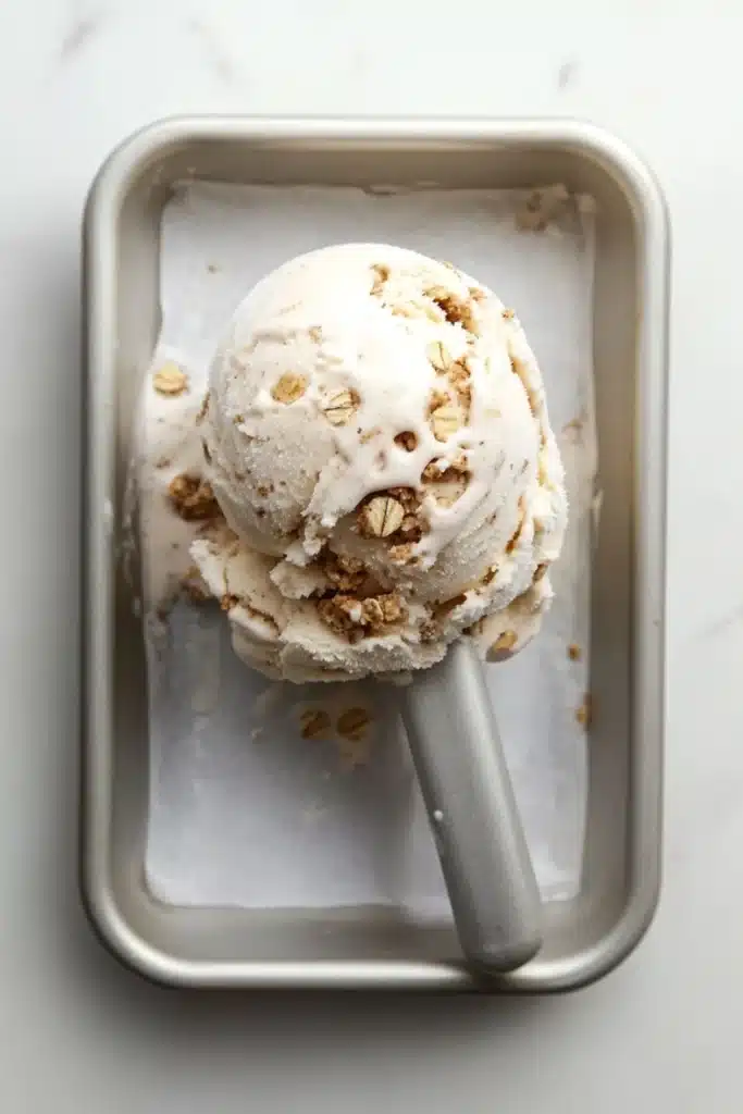 Oatmeal cookie ice cream - the image shows a scoop of vanilla ice cream in a square metal container. the ice cream is topped with chopped nuts, giving it a crunchy texture. the container is sitting on a white marble countertop. a silver ice cream stick is resting on the side of the container.