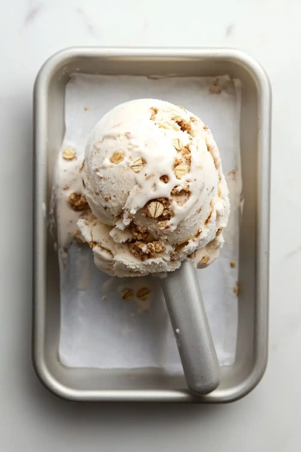 Oatmeal cookie ice cream - the image shows a scoop of vanilla ice cream in a square metal container. the ice cream is topped with chopped nuts, giving it a crunchy texture. the container is sitting on a white marble countertop. a silver ice cream stick is resting on the side of the container.