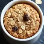 Oatmeal cookie in a mug - the image is a close-up of a round, golden-brown cookie in a white ceramic bowl. the cookie appears to be freshly baked and has a crumbly texture. it is topped with chopped nuts and raisins, giving it a crunchy appearance. the bowl is sitting on a dark, textured surface, and there are scattered nuts around it. the background is blurred, making the cookie the focal point of the image.