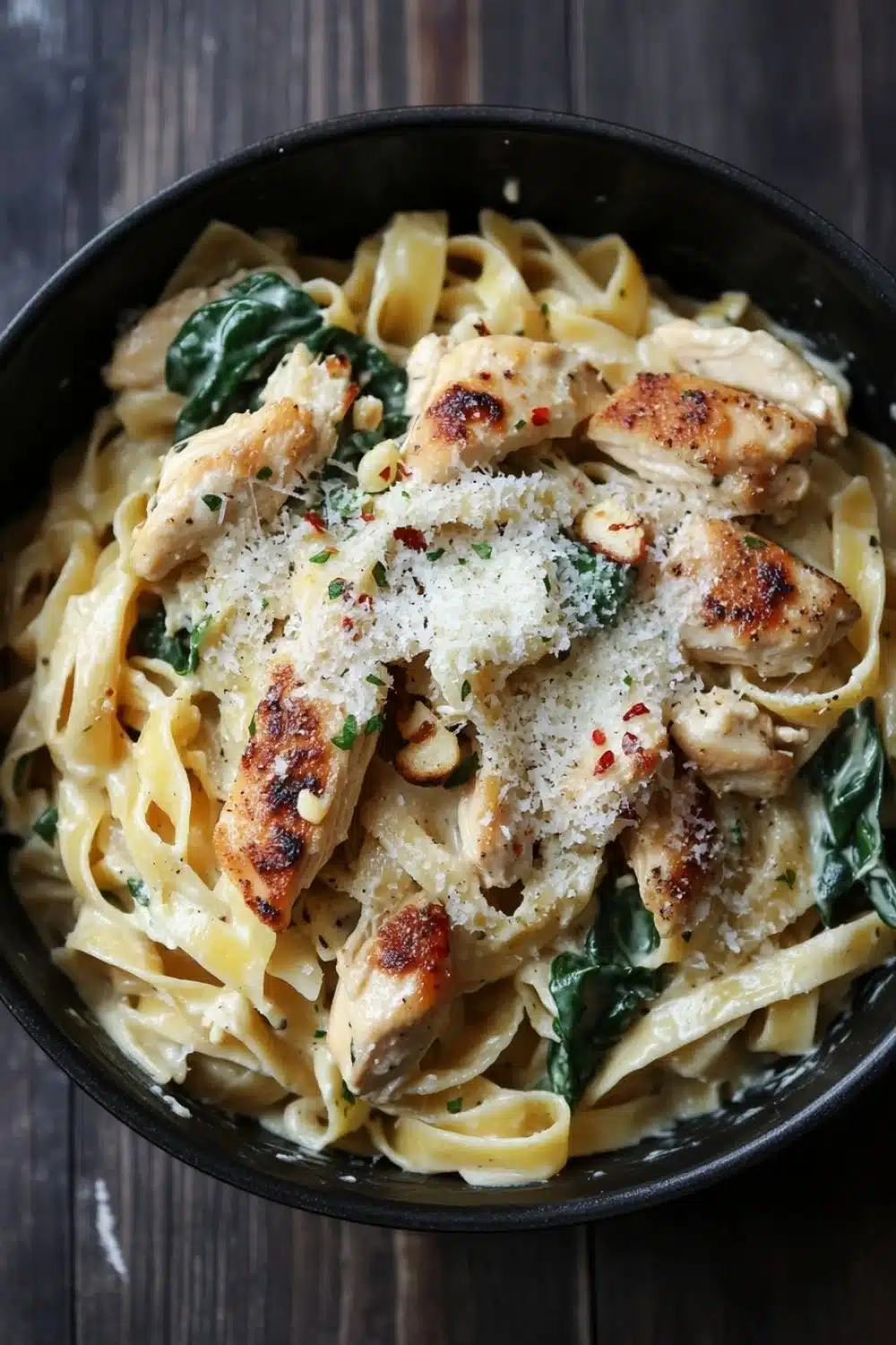 One pot chicken fettuccine - the image is a close-up of a bowl of pasta dish. the pasta appears to be fettuccine alfredo, with chunks of chicken and spinach mixed in. the dish is garnished with grated parmesan cheese and red chili flakes. the bowl is black and is sitting on a wooden table. the background is blurred, but it seems to be a rustic kitchen countertop.