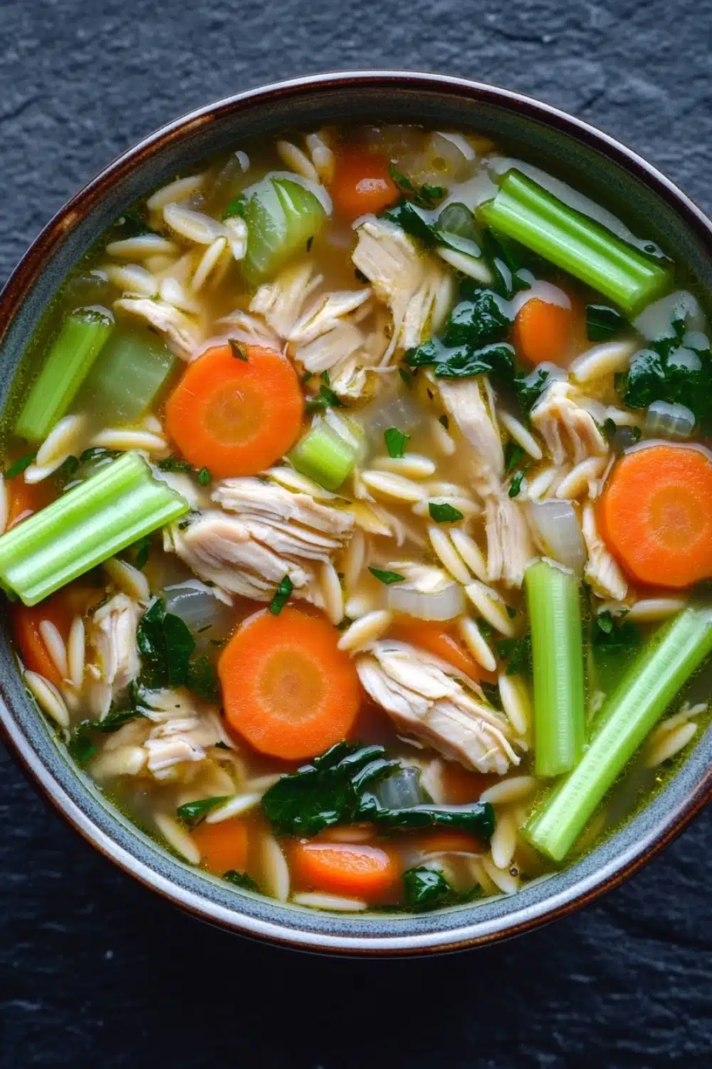 One pot chicken orzo soup - the image is a close-up of a bowl of soup. the soup appears to be a type of noodle soup with chunks of chicken, carrots, celery, and other vegetables mixed in. the broth is a light brown color and looks thick and creamy. the vegetables are bright orange and appear to be fresh and vibrant. the bowl is made of ceramic and is sitting on a dark grey countertop. the background is blurred, making the soup the focal point of the image.