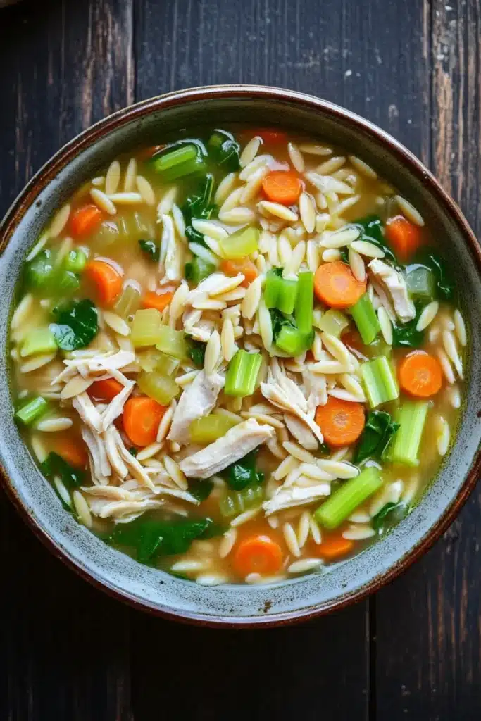 One pot chicken orzo soup - the image is a close-up of a bowl of soup on a wooden table. the soup appears to be a type of noodle soup with chunks of chicken, carrots, celery, and other vegetables mixed in. the bowl is made of ceramic and has a dark brown rim. the broth is a light yellow color and looks thick and creamy. the vegetables are bright orange and green, and they are scattered throughout the soup. the noodles are thin and fluffy, and the chicken is visible in the center of the bowl. the overall appearance of the soup is fresh and appetizing.