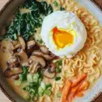 One pot creamy ramen - the image is a close-up of a bowl of ramen soup. the bowl is made of ceramic and has a brown rim. the soup is a creamy yellow color and appears to be thick and creamy. on top of the soup, there is a hard-boiled egg with a runny yolk. the egg is placed on top of a bed of sliced mushrooms and green onions. there are also a few pieces of carrots scattered around the bowl. the background is a wooden table with a beige cloth napkin on the right side.