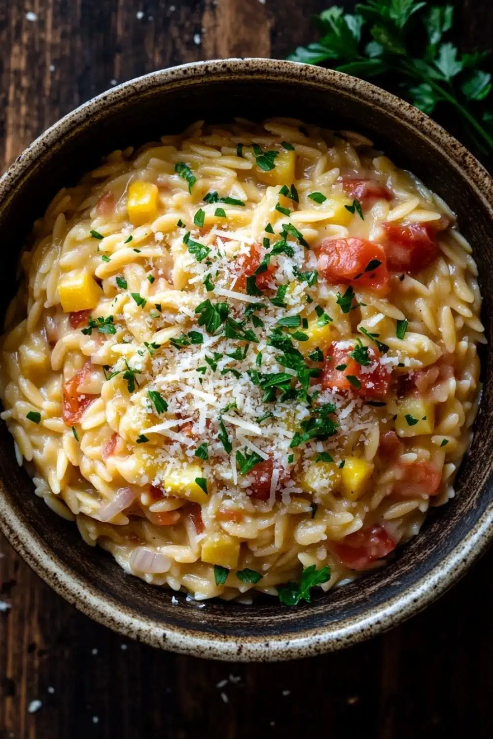 One pot pasta orzo - the image is a close-up of a bowl of pasta dish. the pasta appears to be fusilli, which is a type of italian pasta, and is mixed with diced tomatoes, corn, and other vegetables. the dish is garnished with chopped parsley and grated parmesan cheese. the bowl is made of ceramic and is sitting on a wooden table. there is a sprig of parsley on the side of the bowl. the background is blurred, but it seems to be a rustic kitchen countertop.