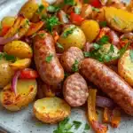 One pot sausage bake - the image shows a plate of food with sausages and potatoes. the plate is white with a speckled pattern and is placed on a wooden table. the sausage is cooked to a golden brown color and is surrounded by diced potatoes, red and yellow bell peppers, and chopped parsley. the potatoes are cut into small cubes and are arranged in a pile on top of the sausage. the dish is garnished with a sprig of parsley, adding a pop of green color to the plate.