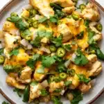 One skillet cheesy green chili chicken - the image is a close-up of a bowl of food. the bowl is made of ceramic and has a light brown color. the food appears to be a dish made with chicken, onions, and jalapeno peppers. the chicken is covered in a yellow sauce, which is drizzled over the top. there are also some chopped cilantro scattered throughout the dish. the dish is garnished with sliced jalapeño peppers and a sprig of cilantro. the background is a white marble countertop.