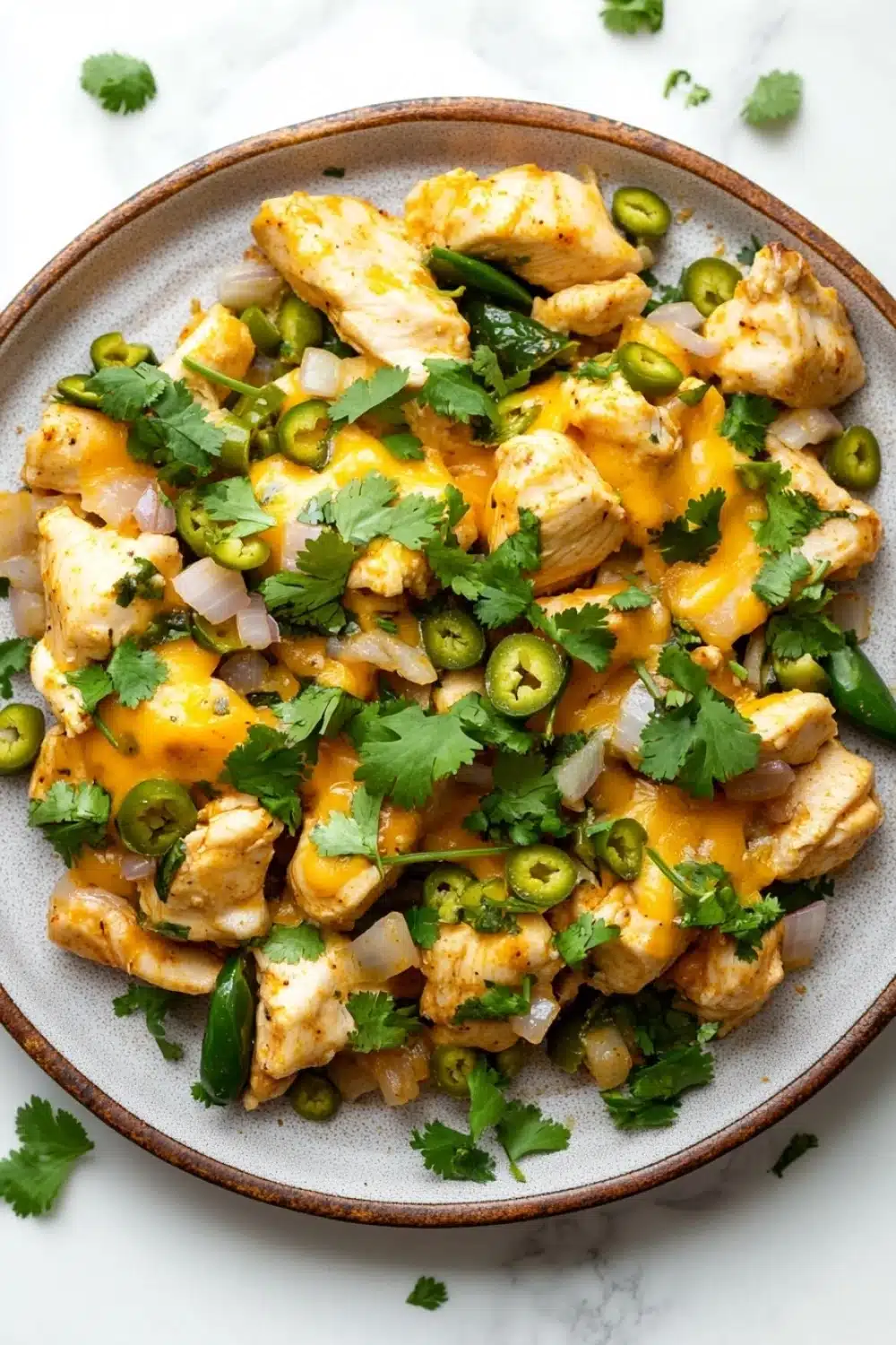 One skillet cheesy green chili chicken - the image is a close-up of a bowl of food. the bowl is made of ceramic and has a light brown color. the food appears to be a dish made with chicken, onions, and jalapeno peppers. the chicken is covered in a yellow sauce, which is drizzled over the top. there are also some chopped cilantro scattered throughout the dish. the dish is garnished with sliced jalapeño peppers and a sprig of cilantro. the background is a white marble countertop.