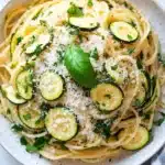 Pasta dinner with zucchini - the image is a close-up of a bowl of spaghetti with zucchini and parmesan cheese. the spaghetti is cooked al dente and is garnished with fresh basil leaves. the bowl is white with a blue and white striped napkin on the side. the background is a white marble countertop. the overall color scheme of the image is warm and inviting.