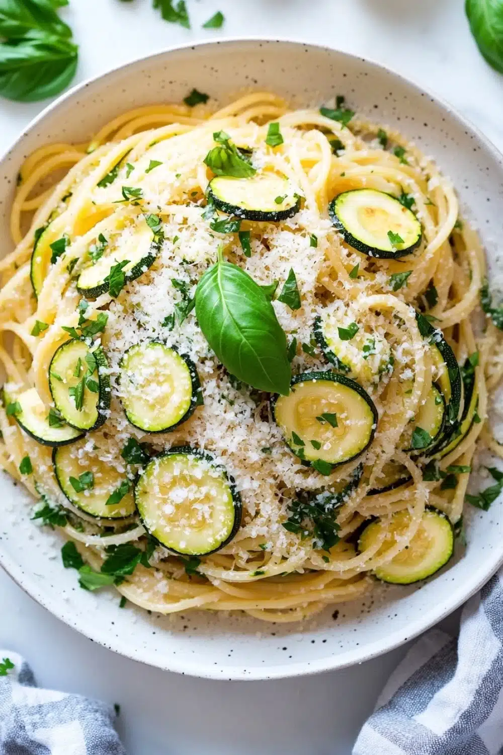 Pasta dinner with zucchini - the image is a close-up of a bowl of spaghetti with zucchini and parmesan cheese. the spaghetti is cooked al dente and is garnished with fresh basil leaves. the bowl is white with a blue and white striped napkin on the side. the background is a white marble countertop. the overall color scheme of the image is warm and inviting.