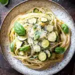 Pasta dinner with zucchini - the image shows a bowl of spaghetti with zucchini and parmesan cheese on top. the spaghetti is cooked al dente and is garnished with fresh basil leaves. the bowl is sitting on a wooden table with a fork and knife on the side. in the background, there is another bowl of grated cheese and a few basil leaves scattered around. the overall color scheme of the image is warm and inviting.