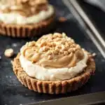 Peanut butter cookie crust dessert - the image shows two small tarts on a black baking tray. the tarts have a golden brown crust and are topped with a generous layer of creamy white cream cheese frosting. the frosting is drizzled with chopped nuts, giving the tarts a crunchy texture. a knife is resting on the tray next to the tartlets. the background is blurred, but it appears to be a kitchen countertop.