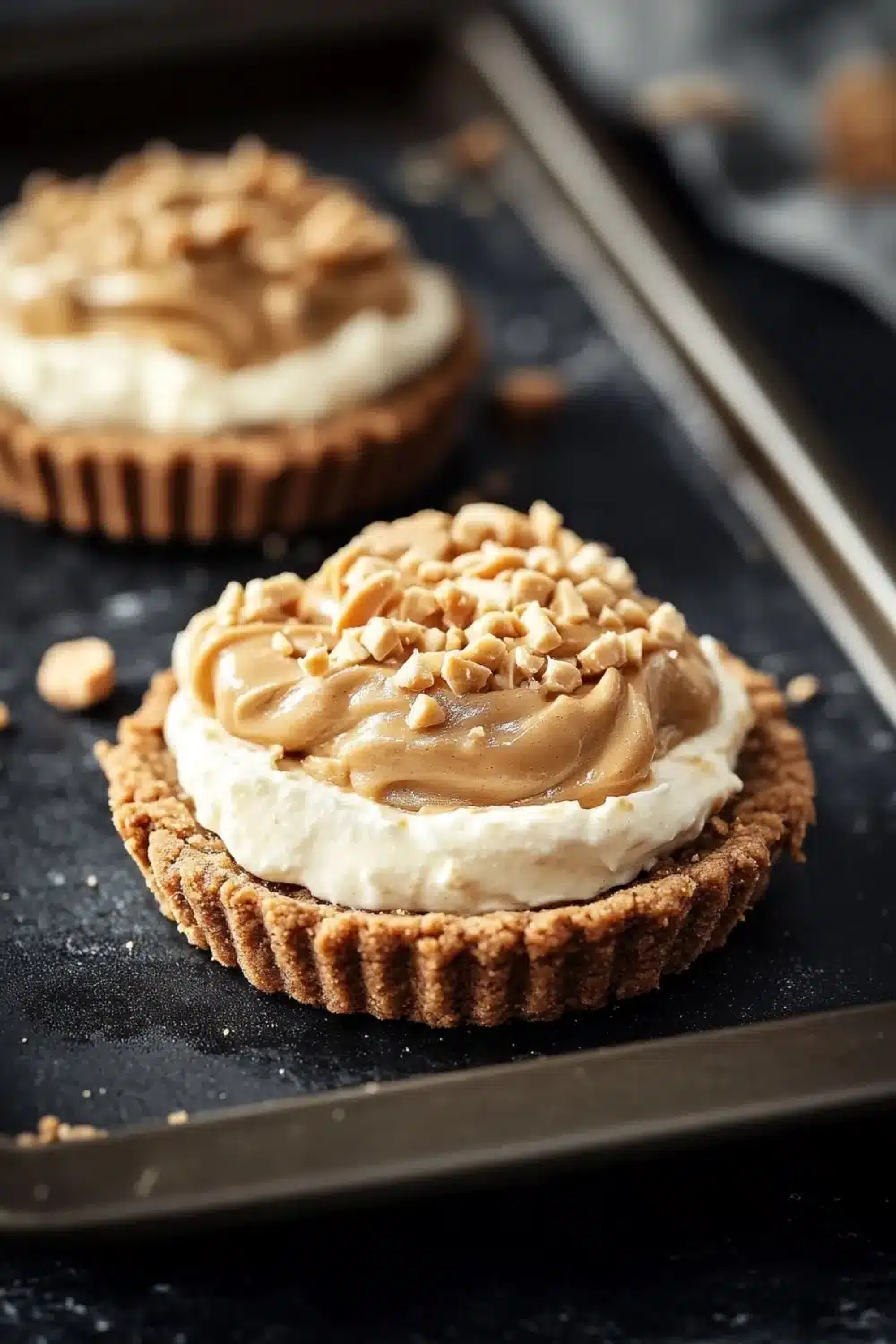 Peanut butter cookie crust dessert - the image shows two small tarts on a black baking tray. the tarts have a golden brown crust and are topped with a generous layer of creamy white cream cheese frosting. the frosting is drizzled with chopped nuts, giving the tarts a crunchy texture. a knife is resting on the tray next to the tartlets. the background is blurred, but it appears to be a kitchen countertop.