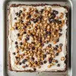 Peanut butter cookie crust dessert - the image shows a rectangular baking tray lined with parchment paper. on top of the parchment paper, there is a layer of white frosting with chocolate chips scattered across it. the frosting appears to be smooth and creamy, and the chocolate chips are dark brown in color. the tray is sitting on a white marble countertop.