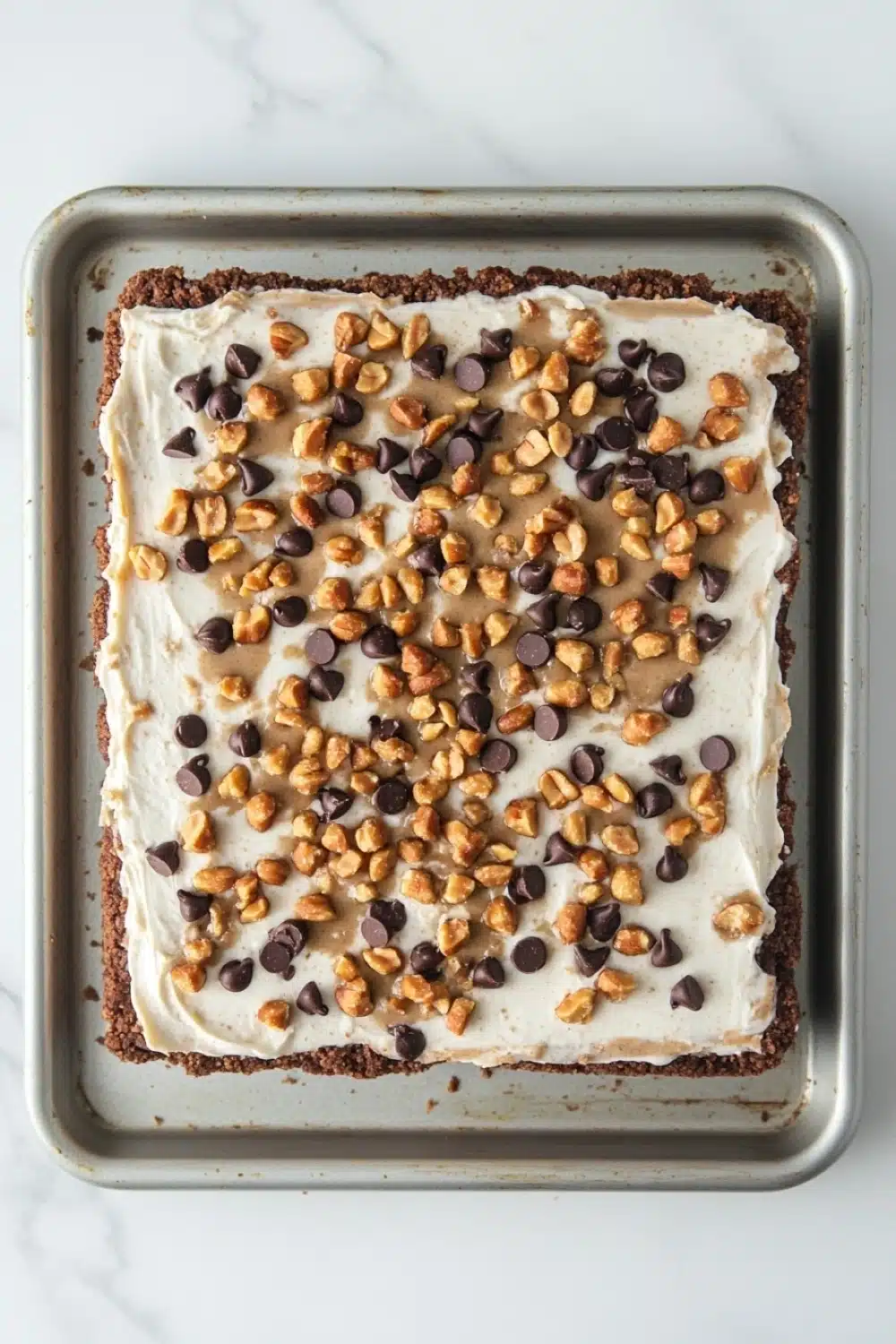 Peanut butter cookie crust dessert - the image shows a rectangular baking tray lined with parchment paper. on top of the parchment paper, there is a layer of white frosting with chocolate chips scattered across it. the frosting appears to be smooth and creamy, and the chocolate chips are dark brown in color. the tray is sitting on a white marble countertop.