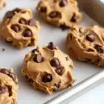Peanut butter cookie dough edible - the image shows a baking tray lined with freshly baked chocolate chip cookies. the cookies are golden brown and have a crumbly texture. they are arranged in a single layer on the tray, with some overlapping each other. the chocolate chips are scattered throughout the cookies, adding a pop of color and texture. the tray is sitting on a white marble countertop.
