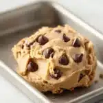 Peanut butter cookie dough edible - the image shows a square metal baking tray with a scoop of peanut butter ice cream in it. the ice cream is a light brown color and has chocolate chips scattered throughout. the tray is placed on a white countertop. the background is blurred, but it appears to be a kitchen countertop with a white sink.