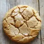 Peanut butter cookie no butter - the image is a close-up of a round cookie on a wooden surface. the cookie appears to be freshly baked and has a golden-brown color. it has a crumbly texture and is covered in a layer of peanut butter. the peanut butter is spread evenly across the cookie, creating a criss-cross pattern. the wooden surface has a weathered and aged appearance, with visible grain and knots. the background is blurred, making the cookie the focal point of the image.