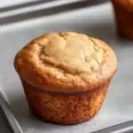Plain yogurt muffin - the image shows two freshly baked muffins on a baking tray. the muffins are golden brown in color and have a crumbly texture on top. they are placed on a white marble countertop. the tray is lined with parchment paper and there are a pair of tongs resting on the tray next to the muffins. the background is blurred, but it appears to be a kitchen countertop with more muffins visible.
