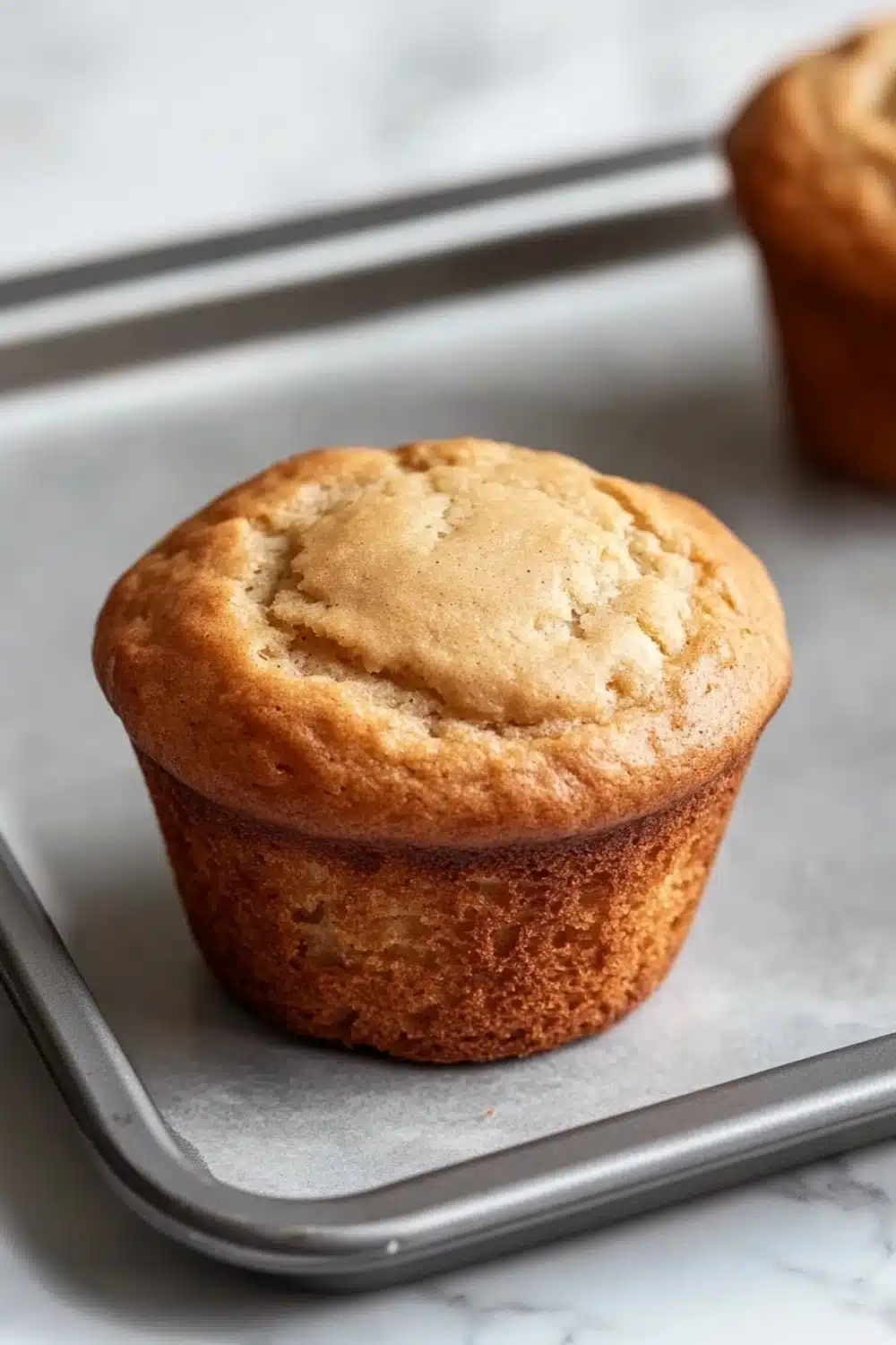 Plain yogurt muffin - the image shows two freshly baked muffins on a baking tray. the muffins are golden brown in color and have a crumbly texture on top. they are placed on a white marble countertop. the tray is lined with parchment paper and there are a pair of tongs resting on the tray next to the muffins. the background is blurred, but it appears to be a kitchen countertop with more muffins visible.