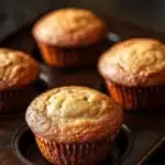 Plain yogurt muffin - the image shows four freshly baked muffins on a wooden tray. the muffins are golden brown in color and have a crumbly texture on top. they are arranged in a row on the tray, with the largest muffin in the center and the two smaller ones on either side. the tray is placed on a dark wooden table with a blurred background. the overall mood of the image is warm and inviting.