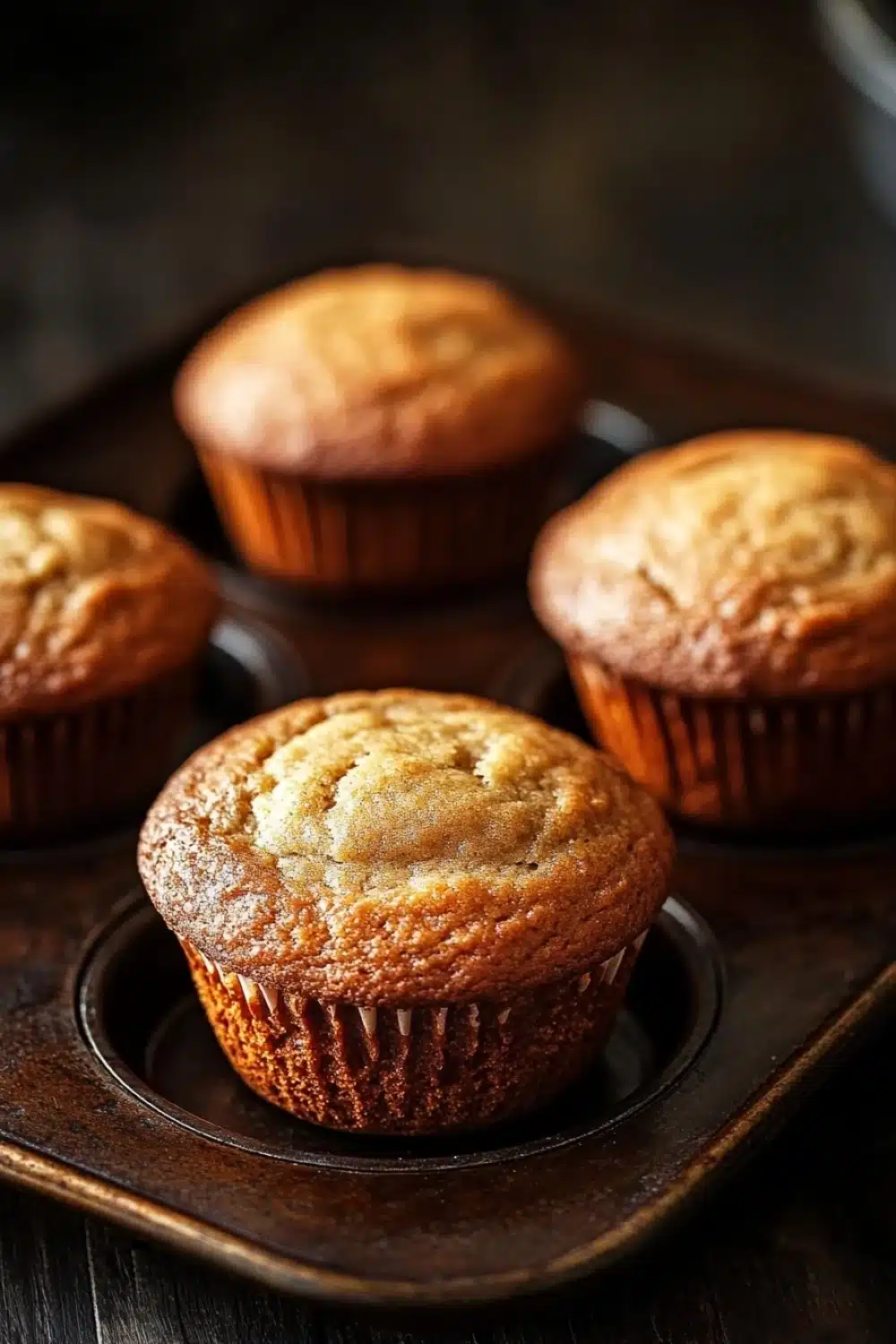 Plain yogurt muffin - the image shows four freshly baked muffins on a wooden tray. the muffins are golden brown in color and have a crumbly texture on top. they are arranged in a row on the tray, with the largest muffin in the center and the two smaller ones on either side. the tray is placed on a dark wooden table with a blurred background. the overall mood of the image is warm and inviting.