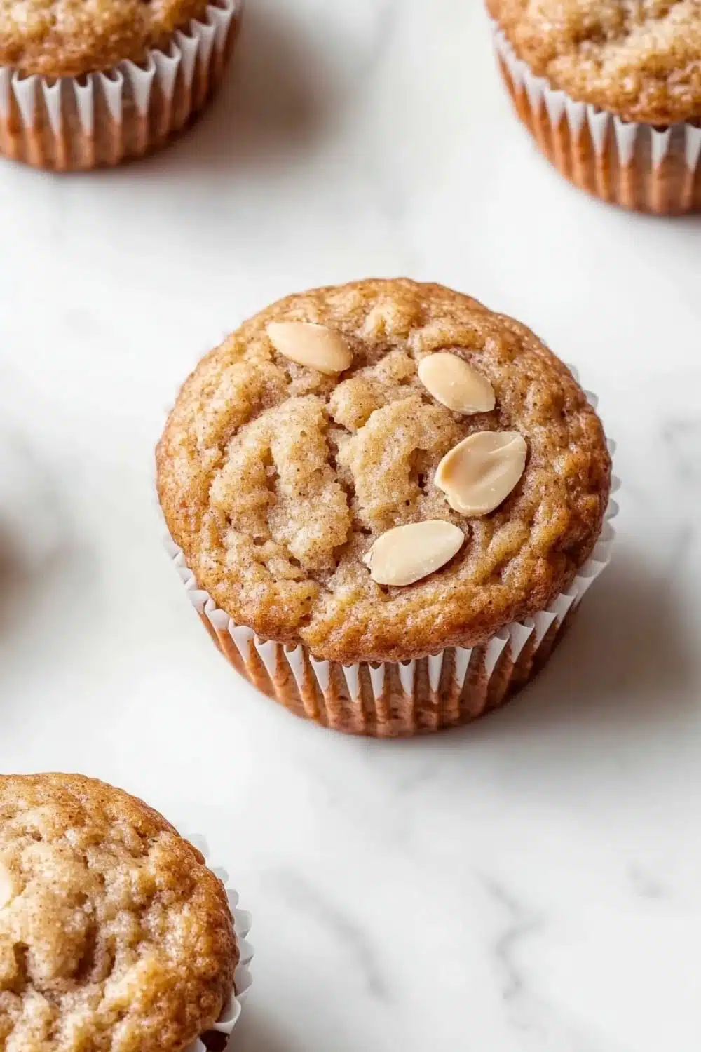 Protein muffin almond flour - the image shows a group of freshly baked muffins on a white marble surface. the muffins are golden brown in color and have a crumbly texture. they are topped with sliced almonds, giving them a crunchy appearance. the background is blurred, making the muffins the focal point of the image.