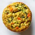 Protein veggie muffin - the image is a close-up of a round muffin on a white marble surface. the muffin appears to be freshly baked and has a golden brown crust. it is filled with a mixture of vegetables, including zucchini, carrots, and spinach, which are visible in the center of the muffin. the vegetables are arranged in a circular pattern, with some overlapping each other. the colors of the vegetables are vibrant and the texture looks soft and fluffy. the background is blurred, making the muffins the focal point of the image.