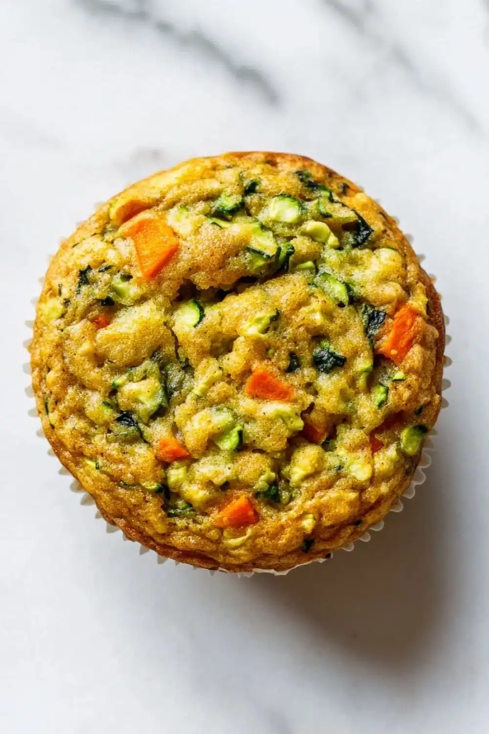 Protein veggie muffin - the image is a close-up of a round muffin on a white marble surface. the muffin appears to be freshly baked and has a golden brown crust. it is filled with a mixture of vegetables, including zucchini, carrots, and spinach, which are visible in the center of the muffin. the vegetables are arranged in a circular pattern, with some overlapping each other. the colors of the vegetables are vibrant and the texture looks soft and fluffy. the background is blurred, making the muffins the focal point of the image.