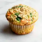 Protein veggie muffin - the image is of a freshly baked muffin on a white marble surface. the muffin is golden brown in color and has a crumbly texture. it is topped with a mixture of chopped vegetables, including spinach, carrots, and zucchini. the vegetables appear to be cooked and have a golden brown color. the top of the muffin has a sprinkle of sesame seeds. the background is blurred, making the muffins the focal point of the image.