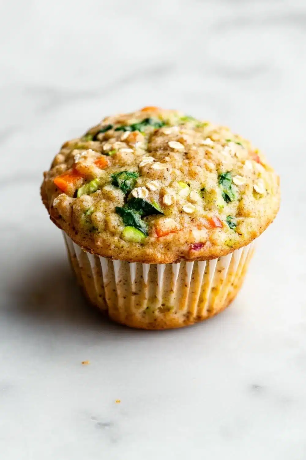 Protein veggie muffin - the image is of a freshly baked muffin on a white marble surface. the muffin is golden brown in color and has a crumbly texture. it is topped with a mixture of chopped vegetables, including spinach, carrots, and zucchini. the vegetables appear to be cooked and have a golden brown color. the top of the muffin has a sprinkle of sesame seeds. the background is blurred, making the muffins the focal point of the image.