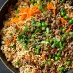 Ramen noodle skillet ground beef - the image is a close-up of a dish in a black bowl. the dish appears to be a type of noodles with ground beef and vegetables mixed in. the noodles are white and fluffy, and the ground beef is a dark brown color. the vegetables include carrots, peas, and green onions. the bowl is filled to the brim with the dish, and it looks like it is ready to be eaten. the background is dark, making the dish stand out.