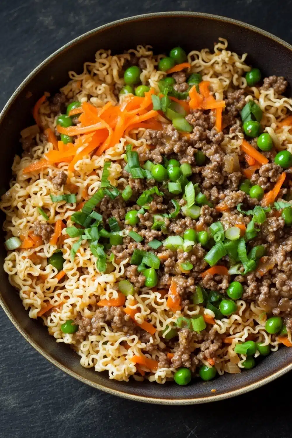Ramen noodle skillet ground beef - the image is a close-up of a dish in a black bowl. the dish appears to be a type of noodles with ground beef and vegetables mixed in. the noodles are white and fluffy, and the ground beef is a dark brown color. the vegetables include carrots, peas, and green onions. the bowl is filled to the brim with the dish, and it looks like it is ready to be eaten. the background is dark, making the dish stand out.