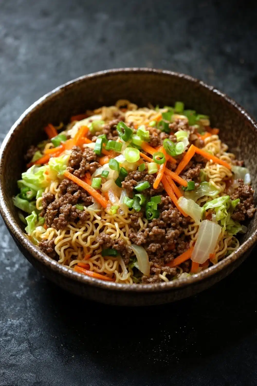 Ramen noodle skillet ground beef - the image is a close-up of a bowl of noodles with minced meat and vegetables. the noodles are yellow and appear to be thin and fluffy. the meat is dark brown and appears to be beef or minced meat. the vegetables are bright orange and are scattered throughout the dish. the dish is garnished with chopped green onions and sliced carrots. the bowl is made of dark brown ceramic and is sitting on a dark grey surface.